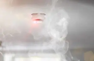 Smoke rising towards a ceiling-mounted smoke detector, which has been activated as indicated by its red warning light.