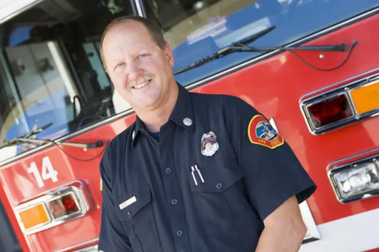 A smiling firefighter in uniform standing in front of a fire truck.