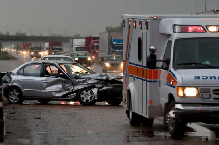 An ambulance arrives at the scene of a serious car accident on a wet road at dusk.